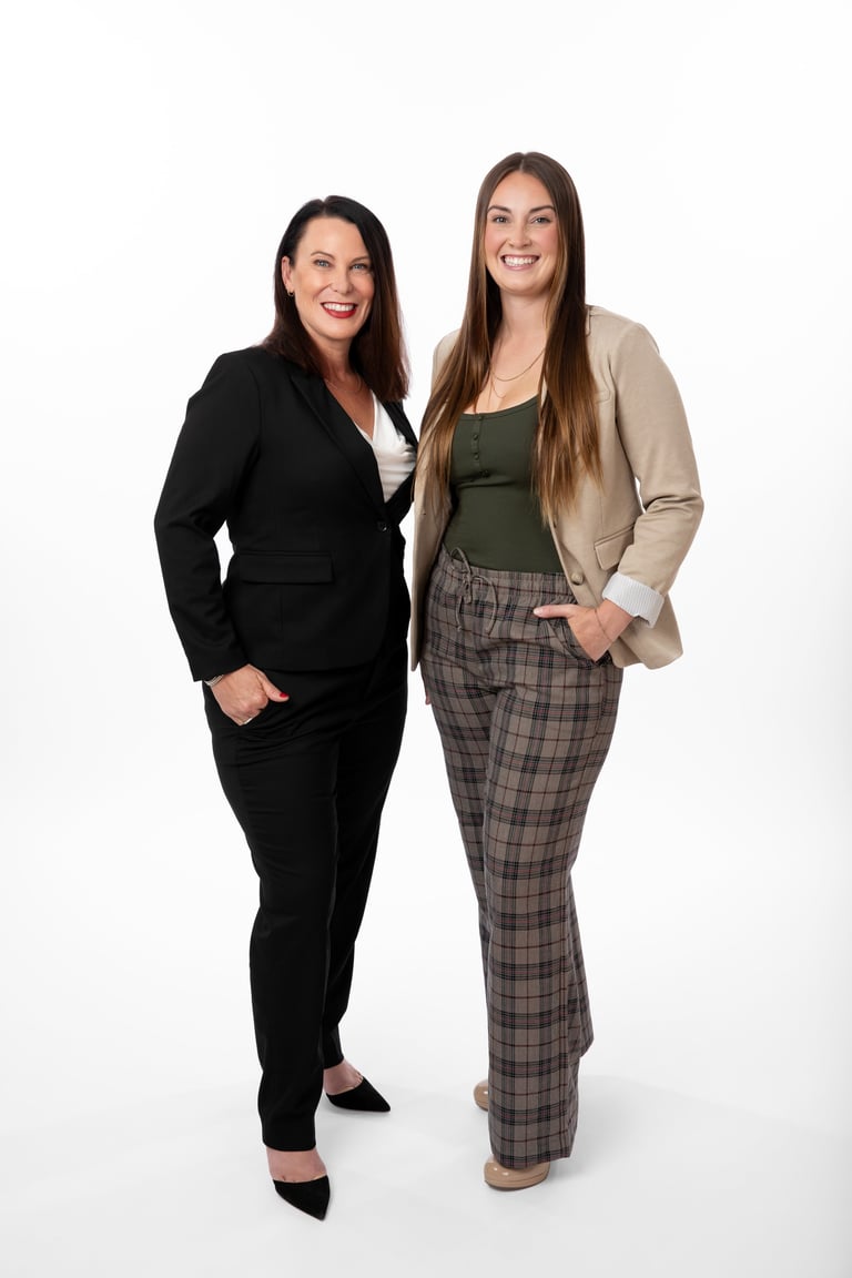Two smiling women standing together against a white background, one in a black blazer and the other in a beige cardigan with plaid pants