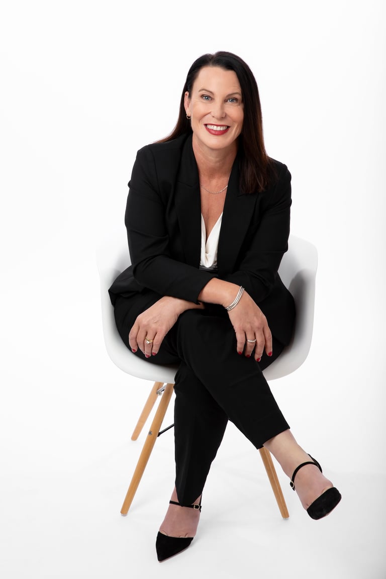 Woman in black business suit sitting on white chair, smiling at camera against white background