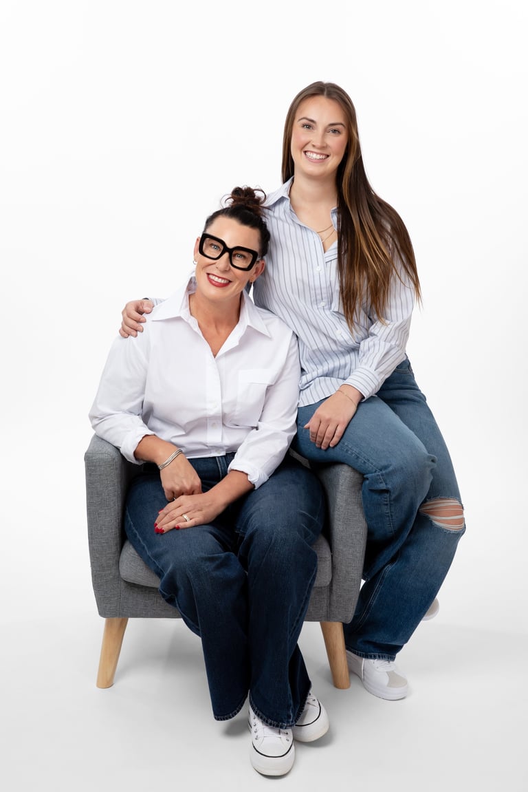 Two smiling women posing together on a gray chair against a white background. One wearing glasses and white shirt, the other in blue striped shirt.